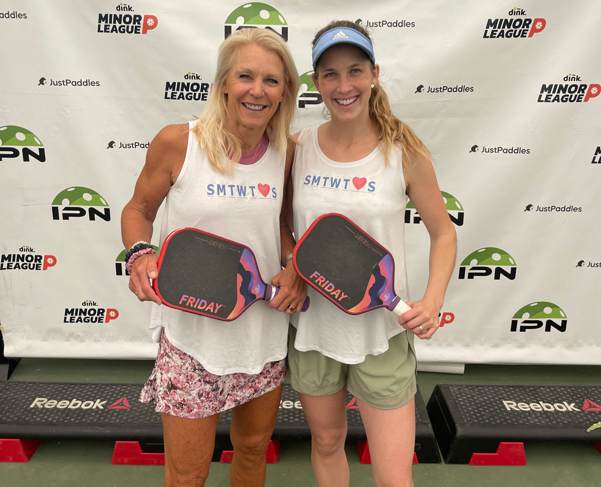 Two women holding pickleball paddles, standing in front of a sports event backdrop.