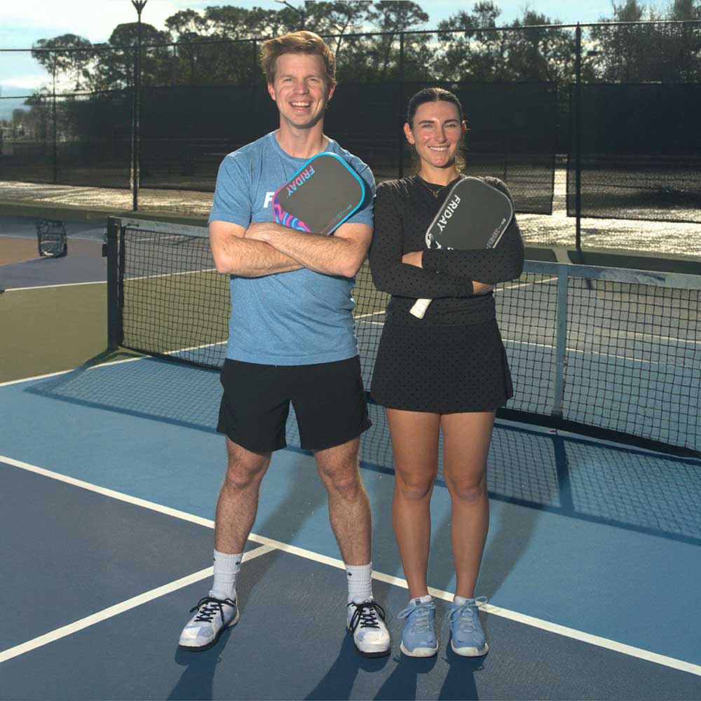 A man and a woman stand on a blue pickleball court, smiling and holding paddles.