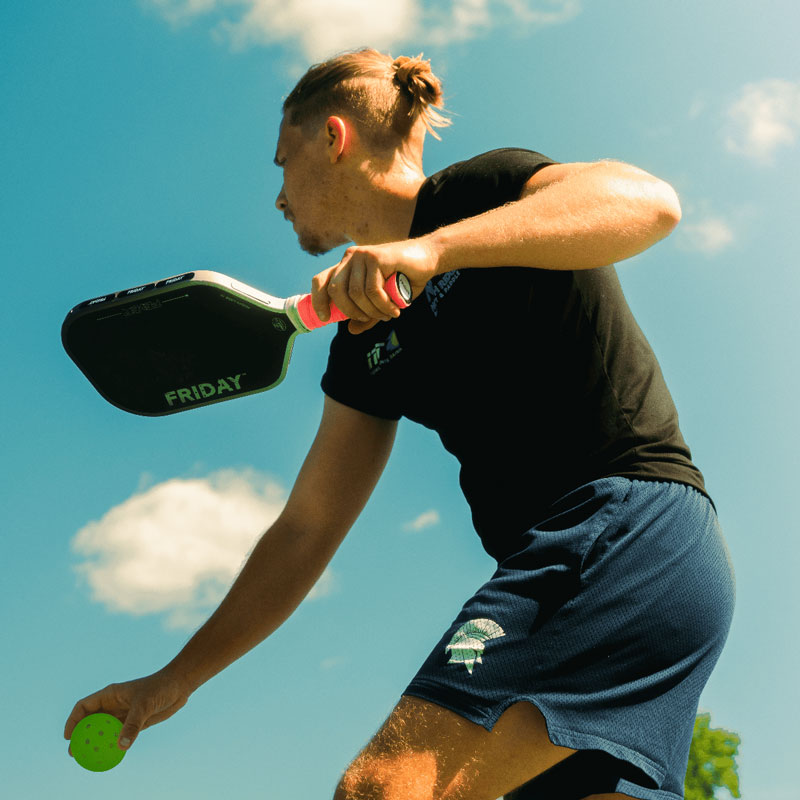 Person playing pickleball outdoors with a paddle and ball under blue sky.