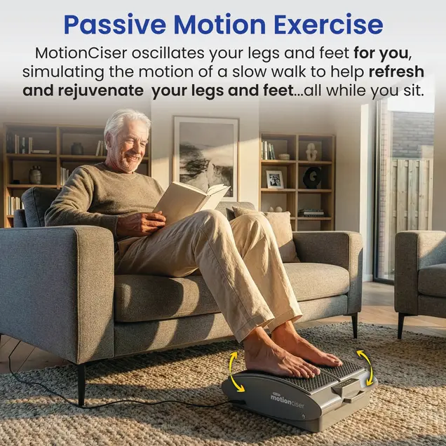 An older man sits on a sofa reading a book with his feet on a passive motion exercise machine.