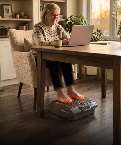 A woman sits at a desk using a laptop while her feet rest on a moving motionciser machine.