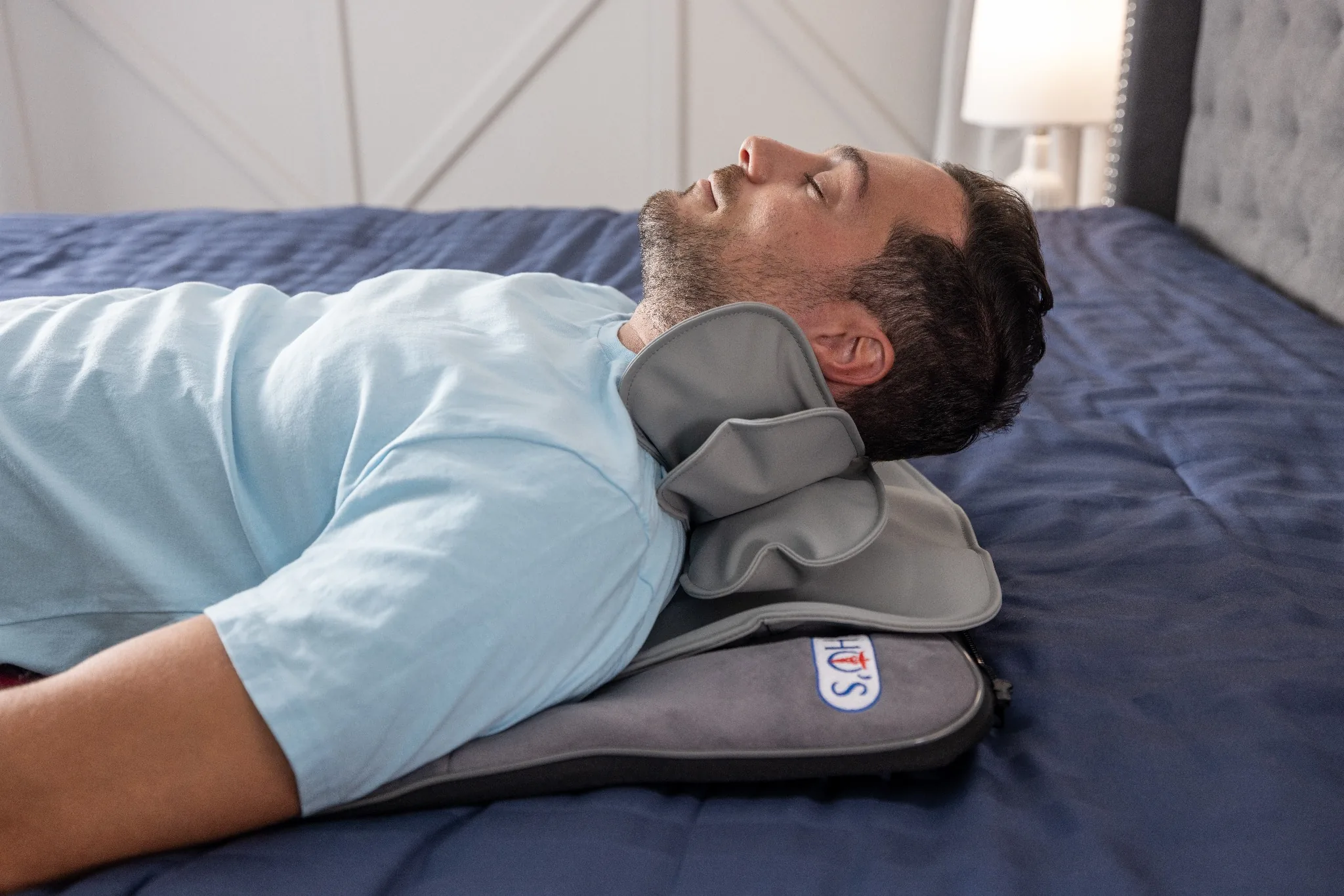 A man lies on his back in bed, his head resting on a gray therapeutic neck pillow.
