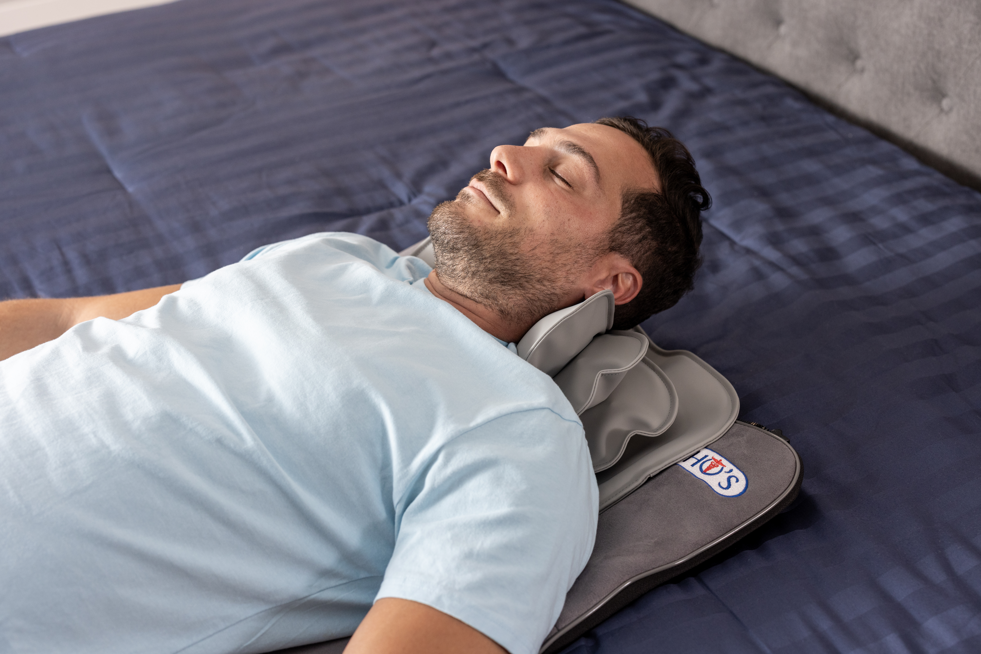 Man lying on bed with a neck pillow, eyes closed, wearing a light blue shirt.