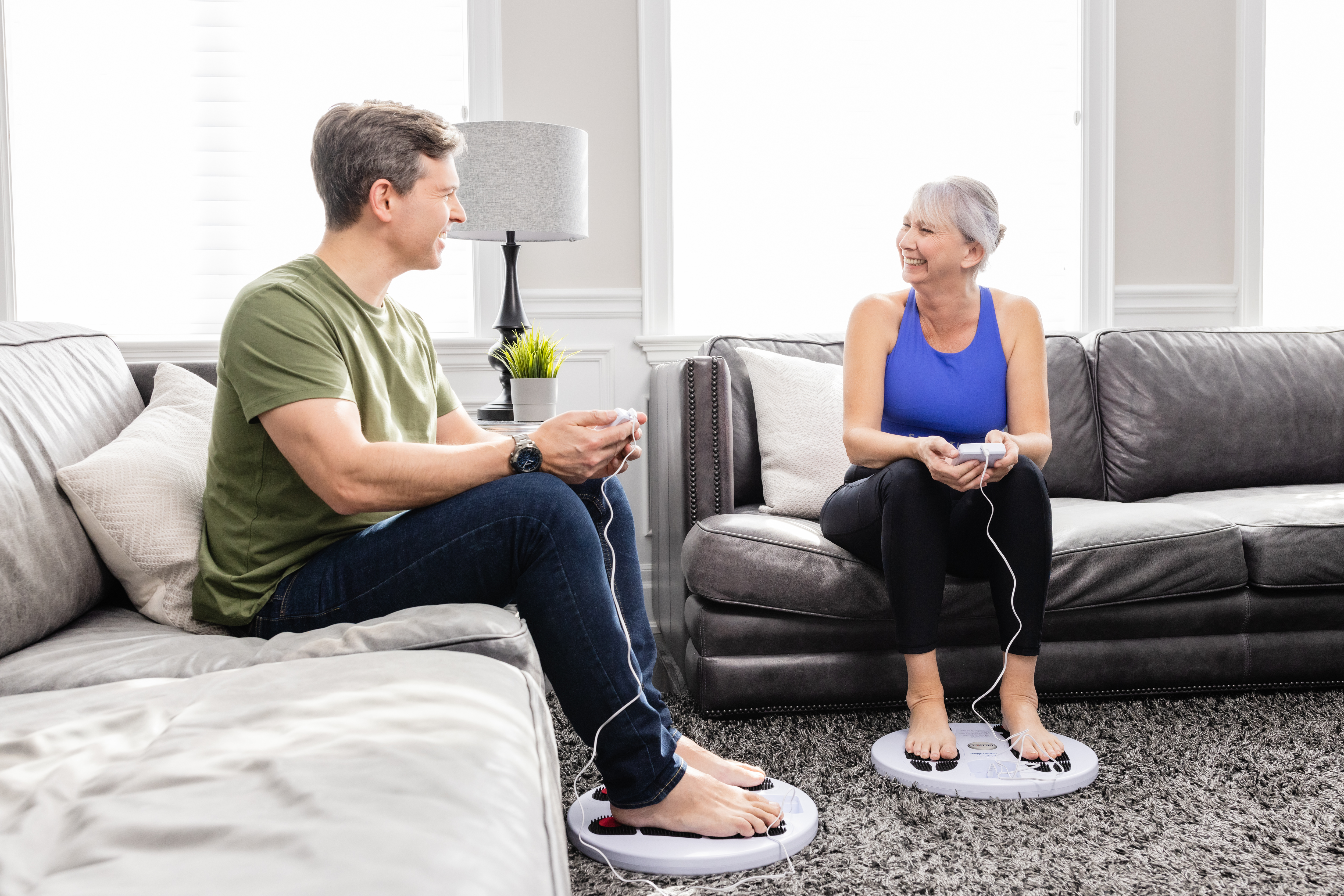 Two people sitting on couches using foot massagers and smiling at each other.