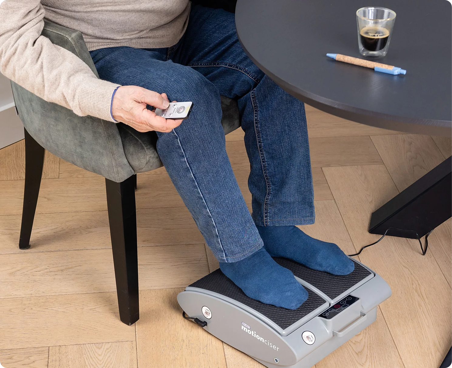 Person using a foot exercise machine under a table, holding a remote control.