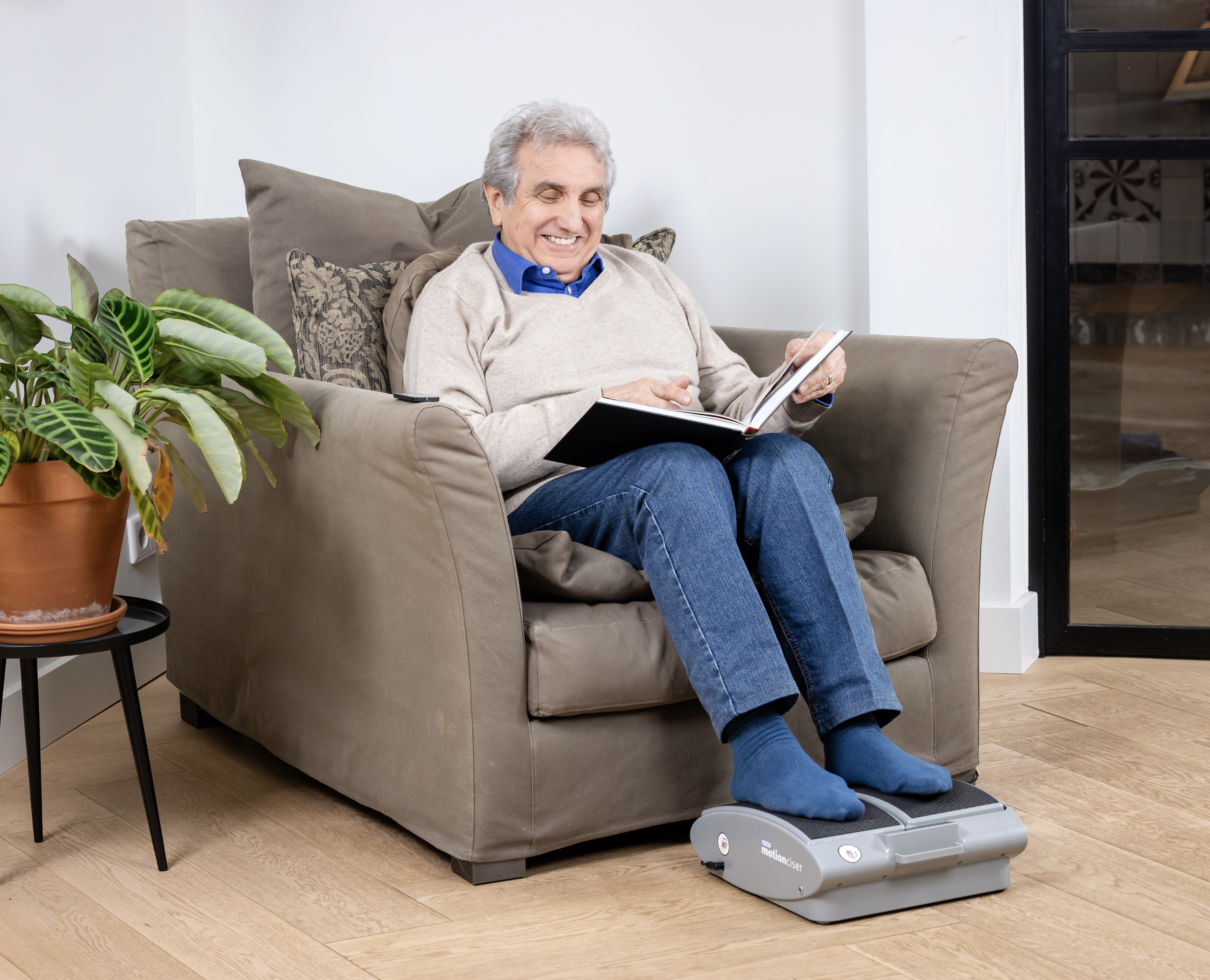 Smiling person sitting in a chair, reading a book, with feet on a foot massager.