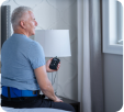 Older man sitting on a bed holding a remote control, looking out the window.