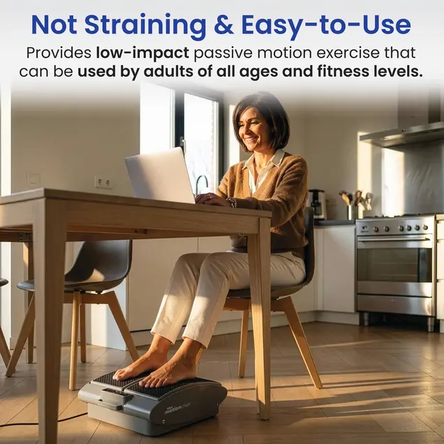 A woman uses a laptop at a table while her feet rest on a passive leg exercise machine.