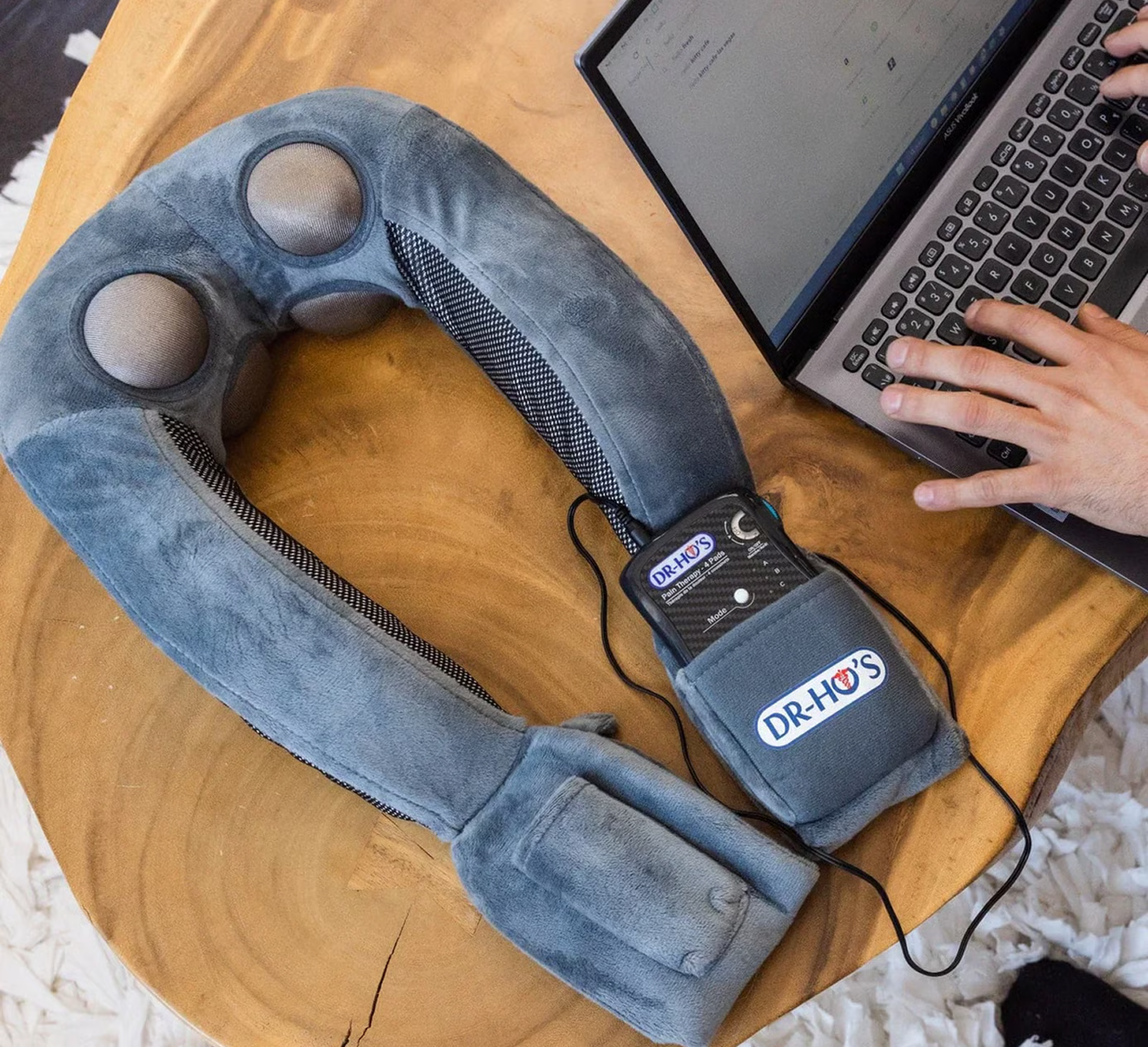 Gray neck massager on a wooden table near a person using a laptop.
