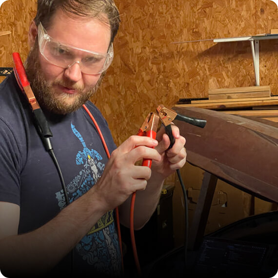 Man wearing safety goggles holding jumper cables in a workshop.