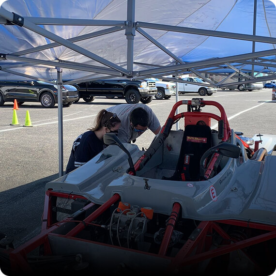 Two people examining a race car under a canopy in a parking lot.