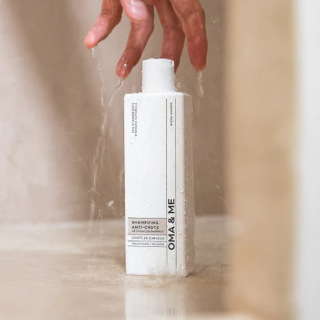 A hand holds a white bottle of OMA & ME shampoo under running water in a shower.