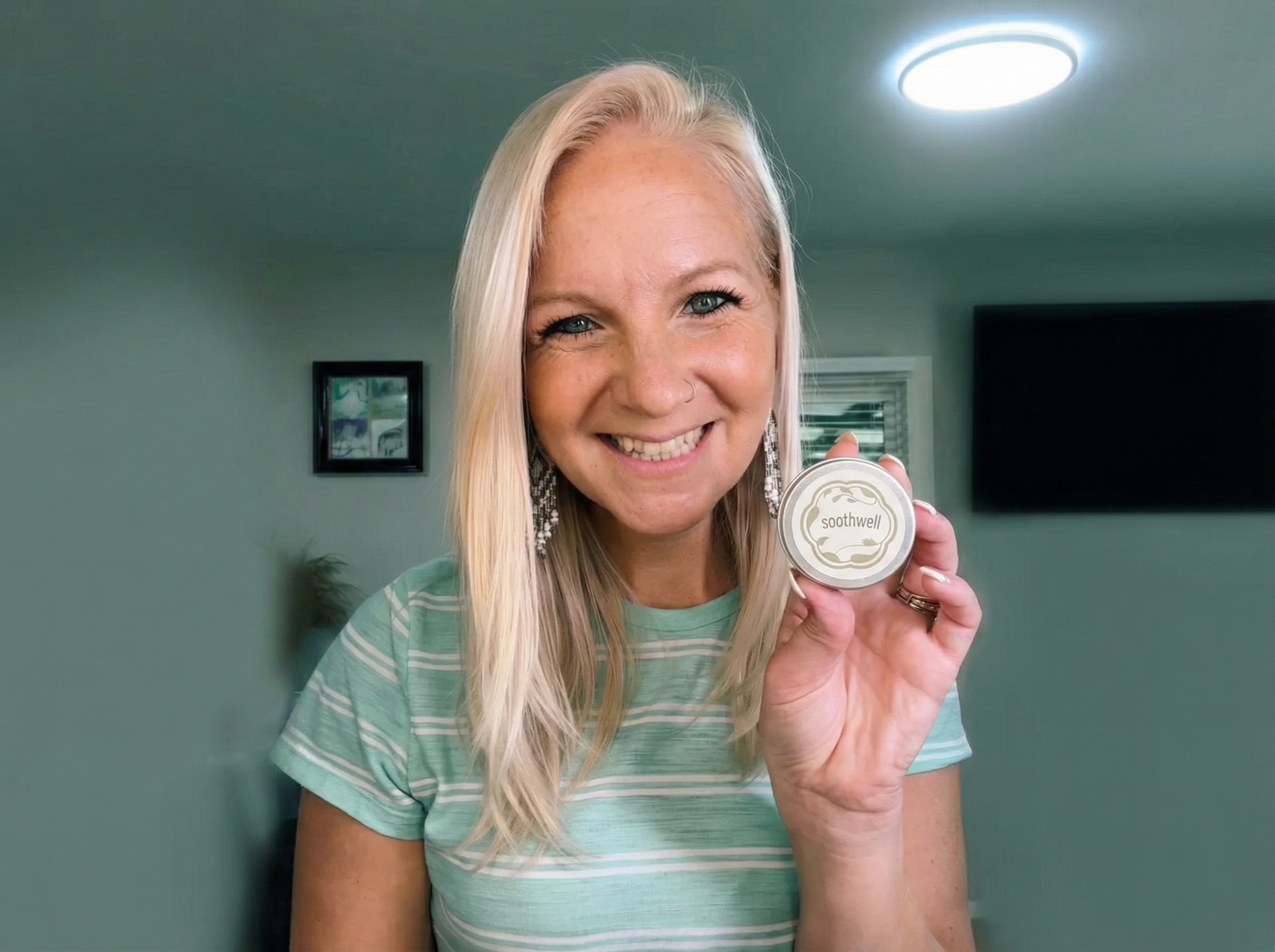 A smiling blonde woman in a striped t-shirt holds up a small tin of Soothwell.