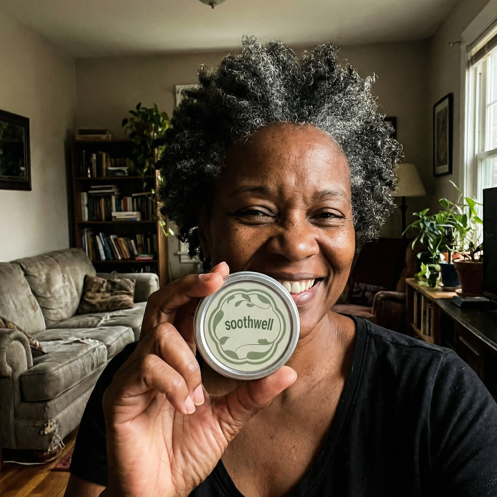 A smiling woman with curly grey hair holds up a small round tin labeled 'soothwell' in her living room.