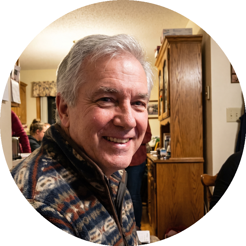 A smiling, grey-haired man in a patterned fleece jacket looks at the camera from inside a house.