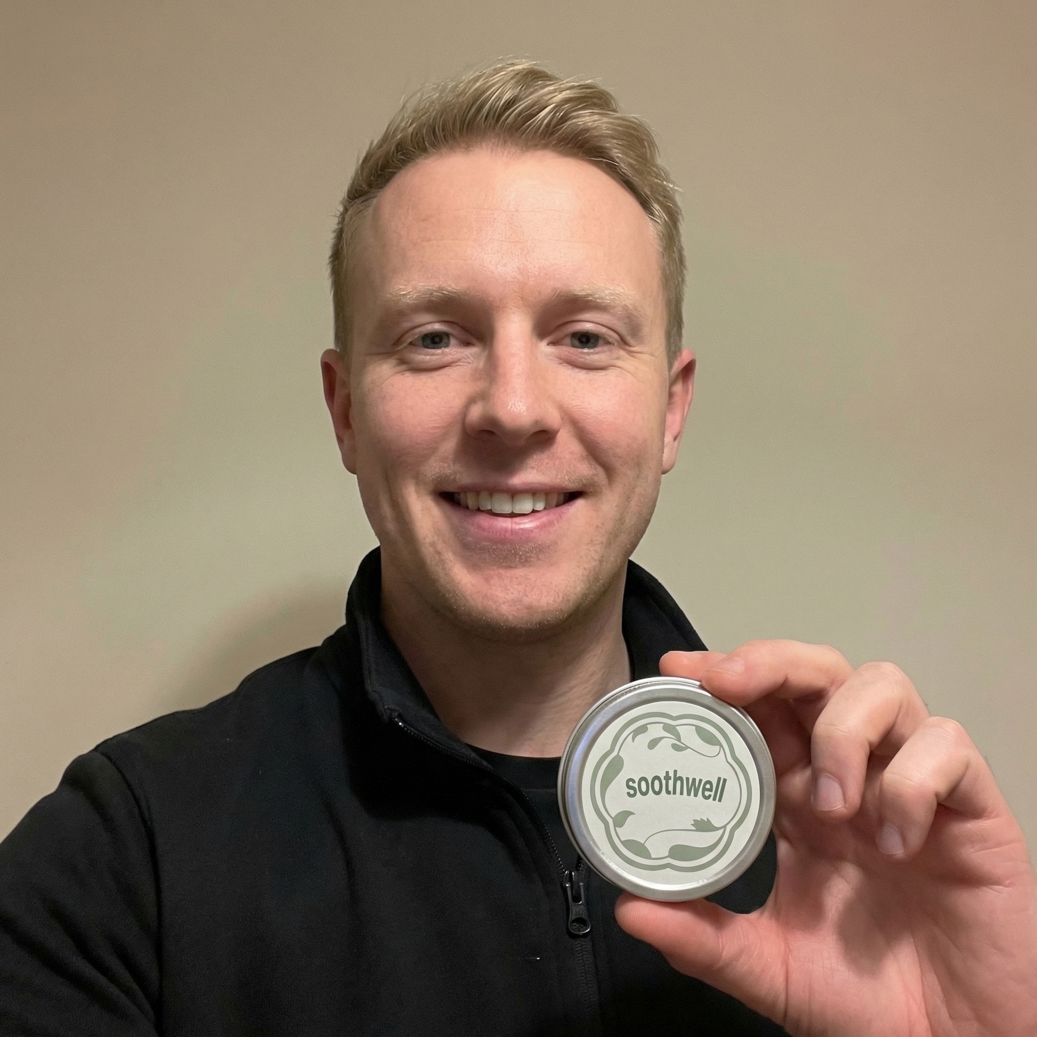 A smiling man with blond hair holds up a round silver tin with the word 'soothwell' on it.