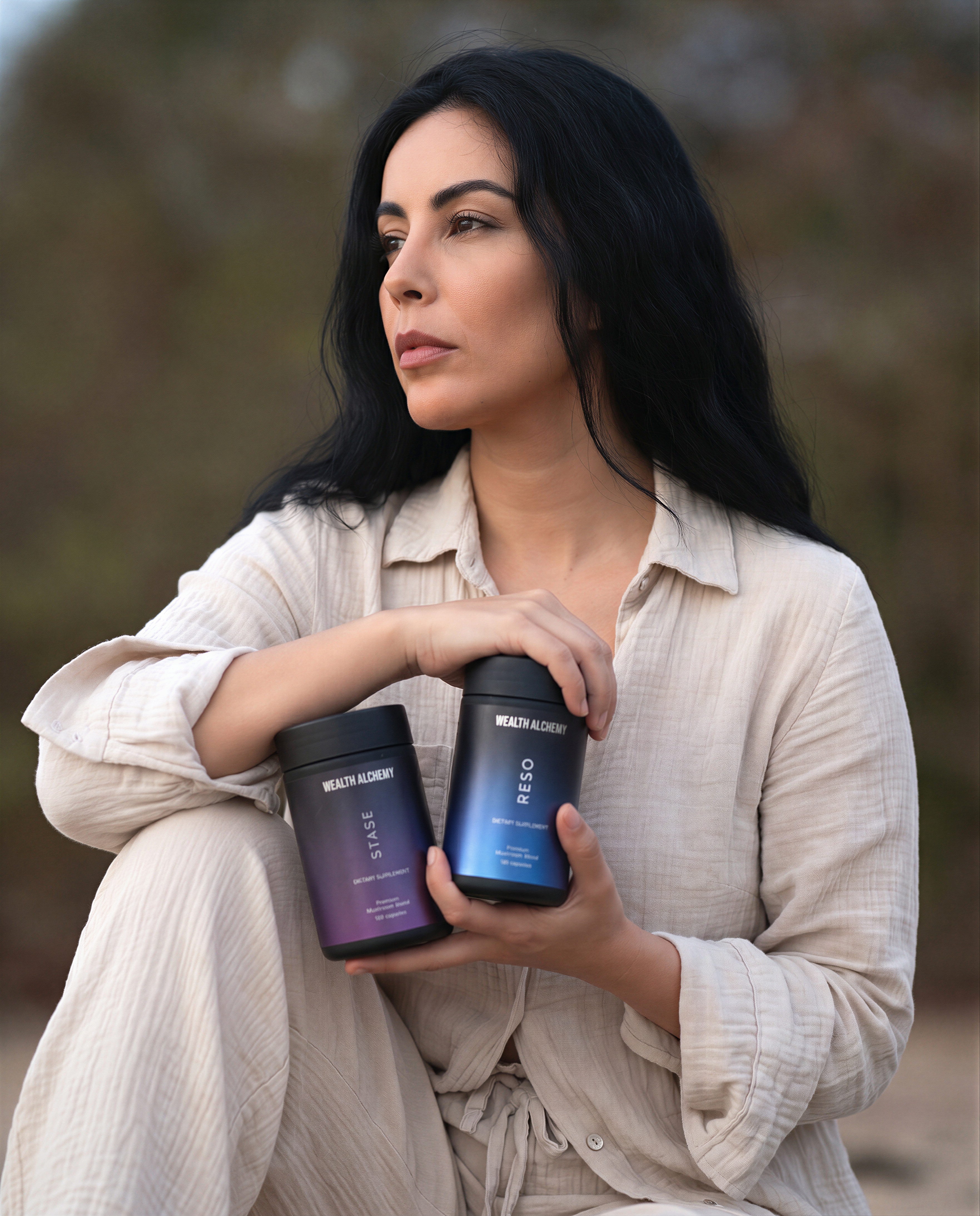 A woman with long dark hair sits outside, holding two supplement containers from the brand Wealth Alchemy.