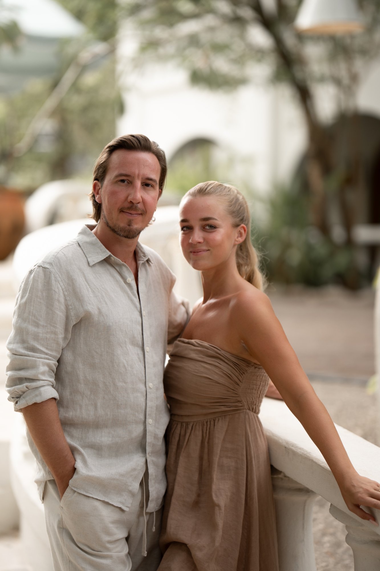A man and a woman in neutral-toned clothes stand together on a balcony, looking at the camera.