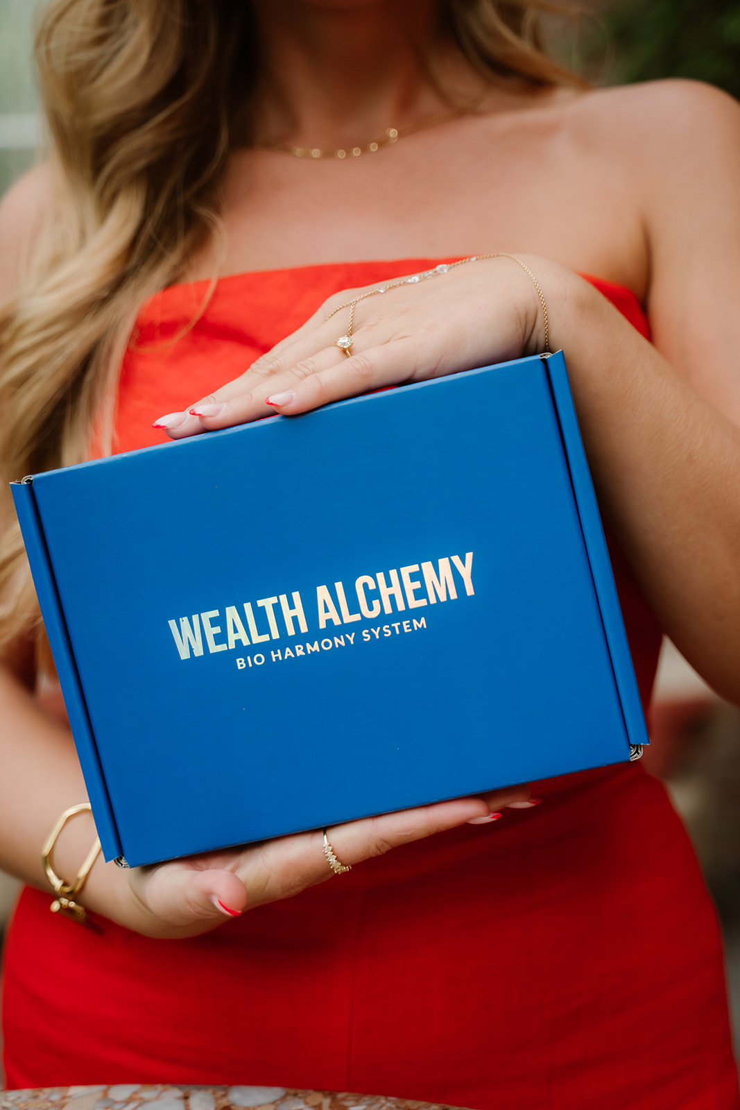 A woman in a red top holds a blue box that reads 'Wealth Alchemy Bio Harmony System'.