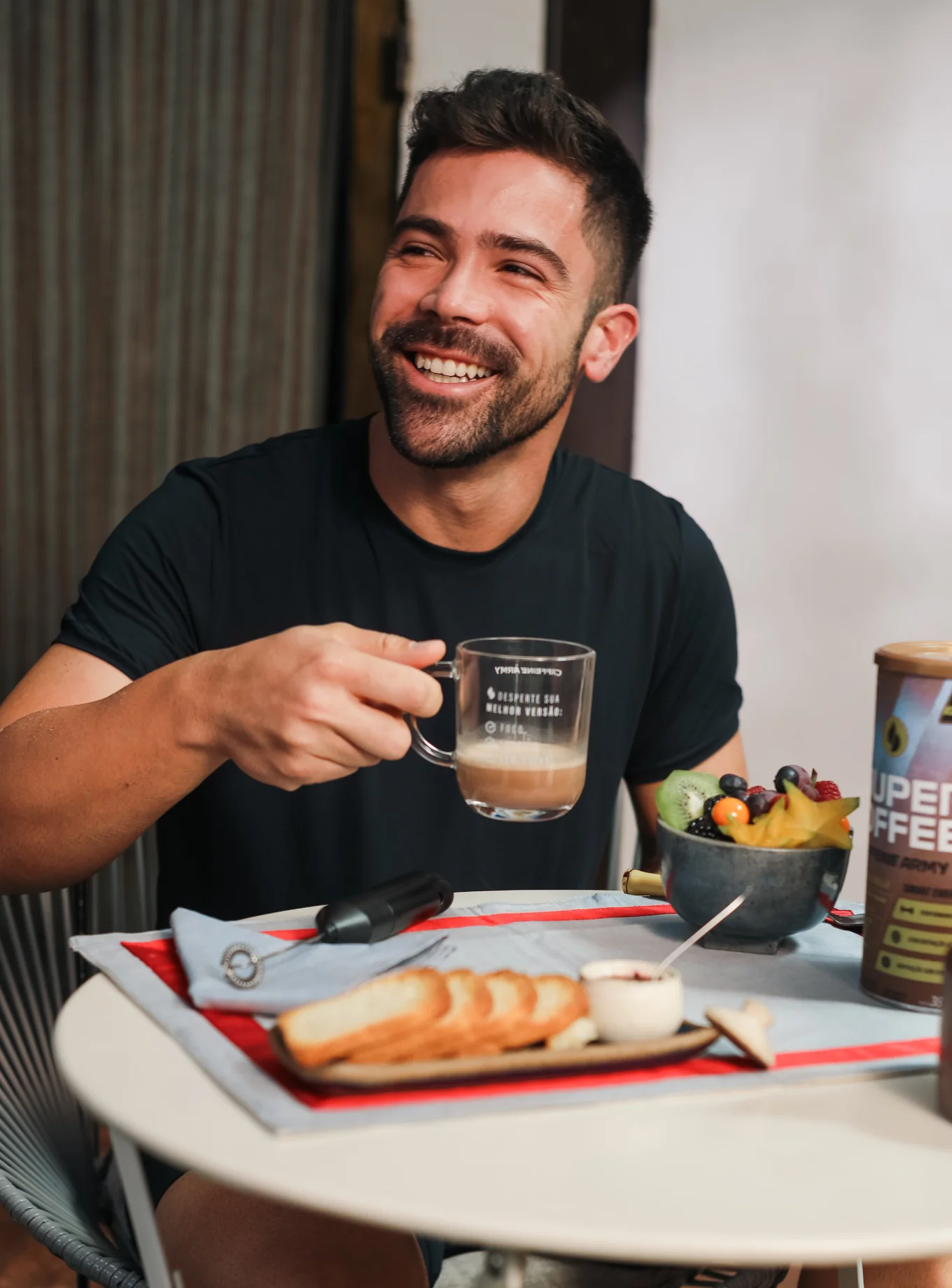 Man smiling while holding a coffee mug, with fruit and bread on the table.