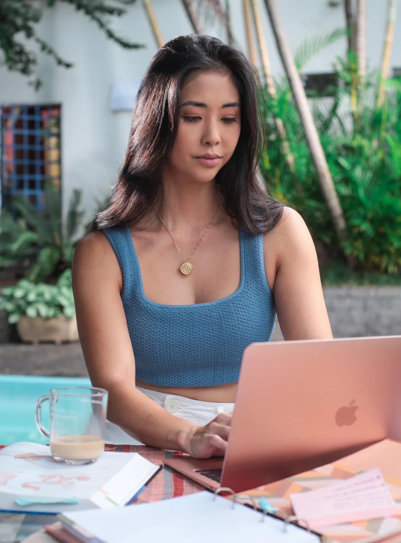 Woman working on a laptop by a pool with notebooks and coffee.