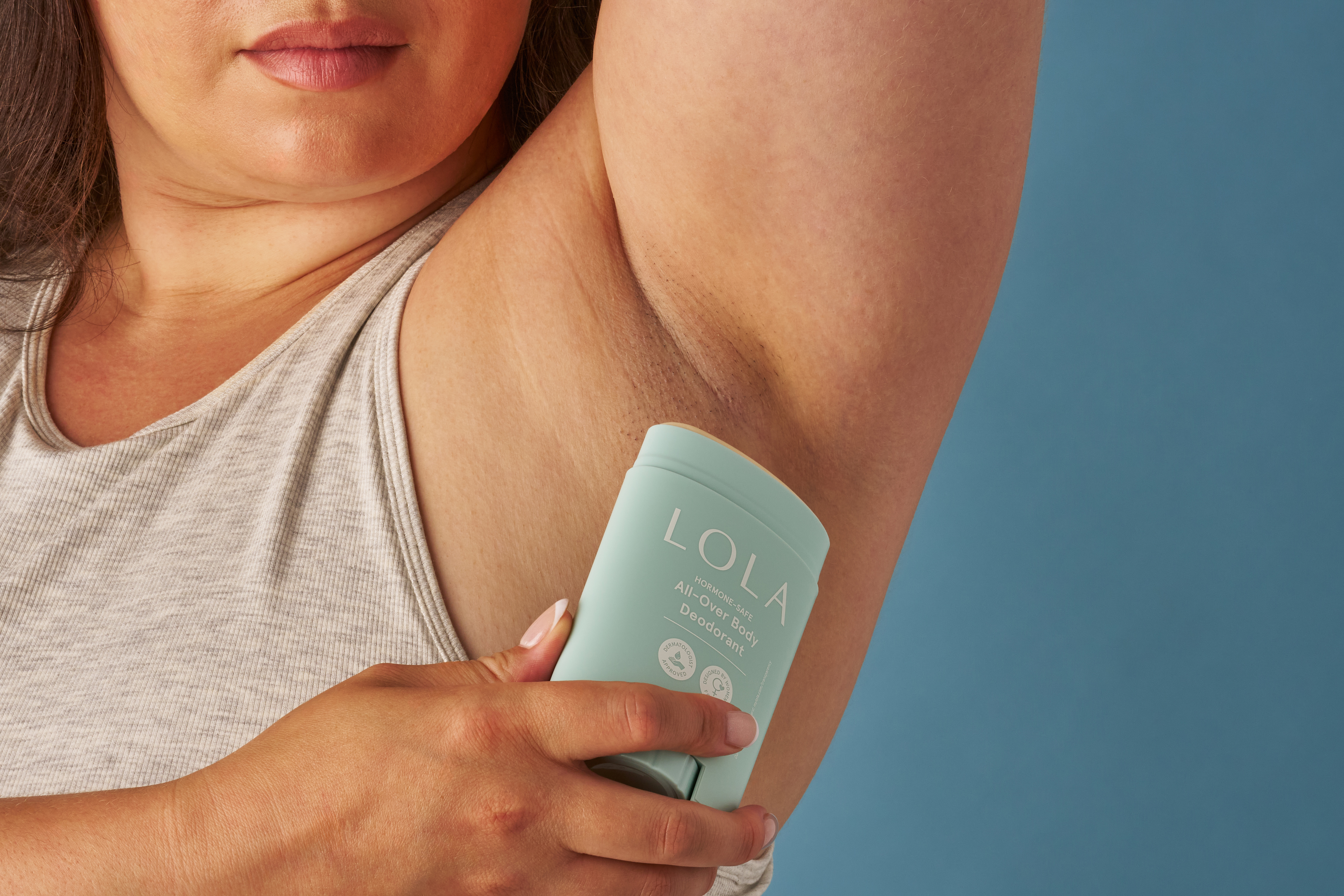 A person in a grey tank top applies Lola all-over body deodorant to their underarm.