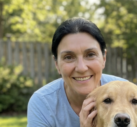 A smiling woman with dark hair, wearing a blue shirt, poses with a golden retriever outdoors.