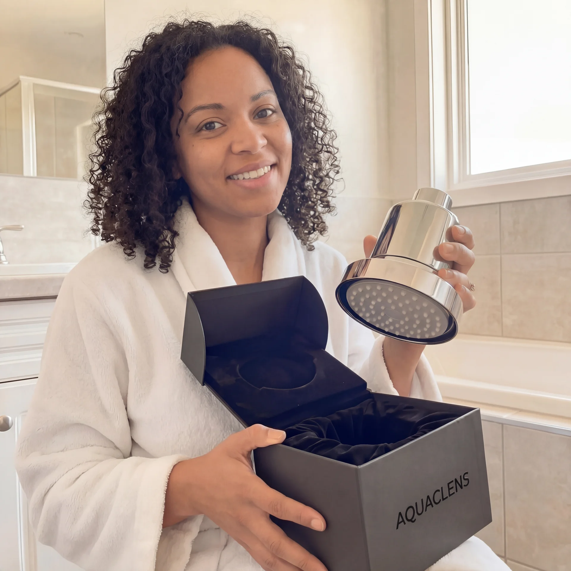 A smiling woman in a white bathrobe unboxes a new chrome shower head in a bathroom.