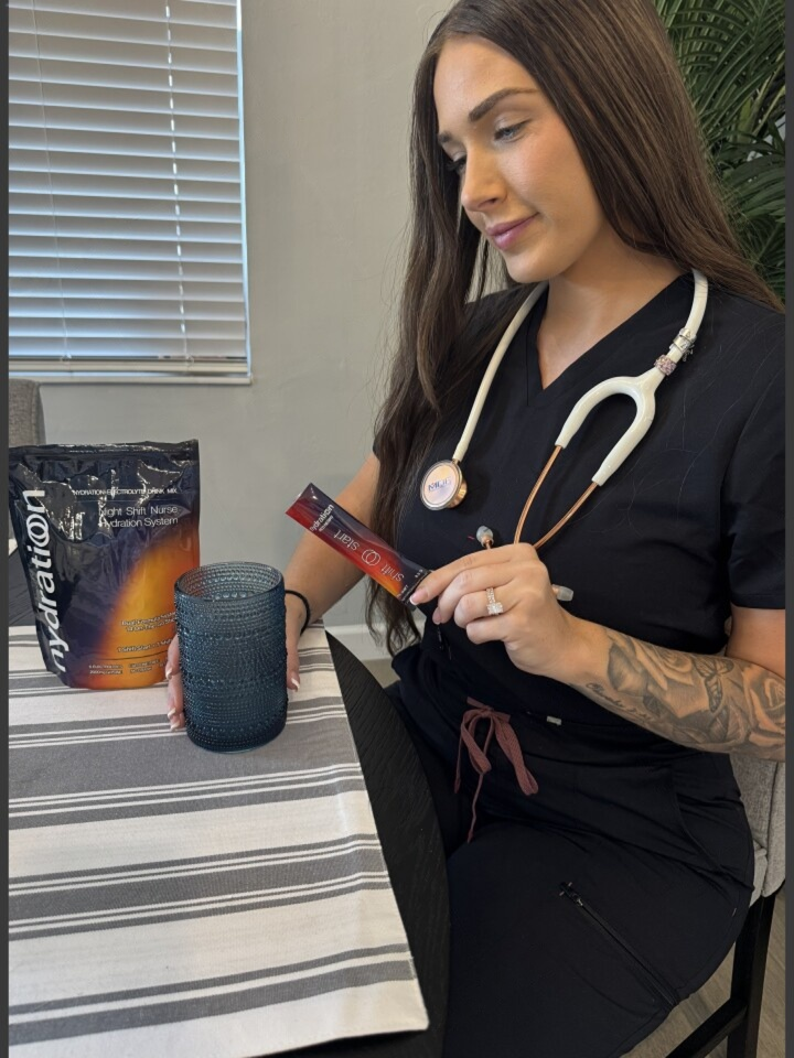 A woman in scrubs with a stethoscope holds a hydration packet over a glass at a table.