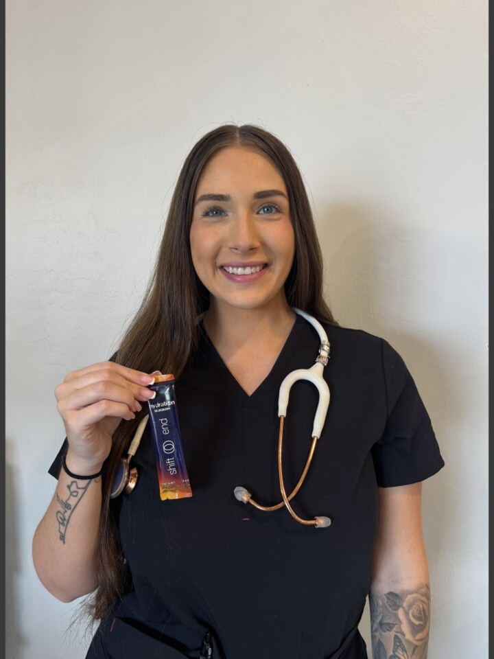 A smiling woman in black scrubs with a stethoscope holds a hydration packet in front of a white wall.