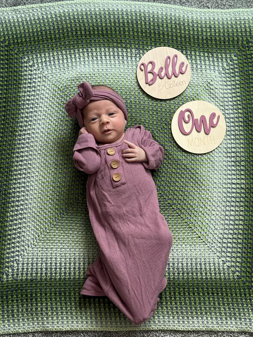 Newborn in purple outfit on green blanket with wooden name and age markers.