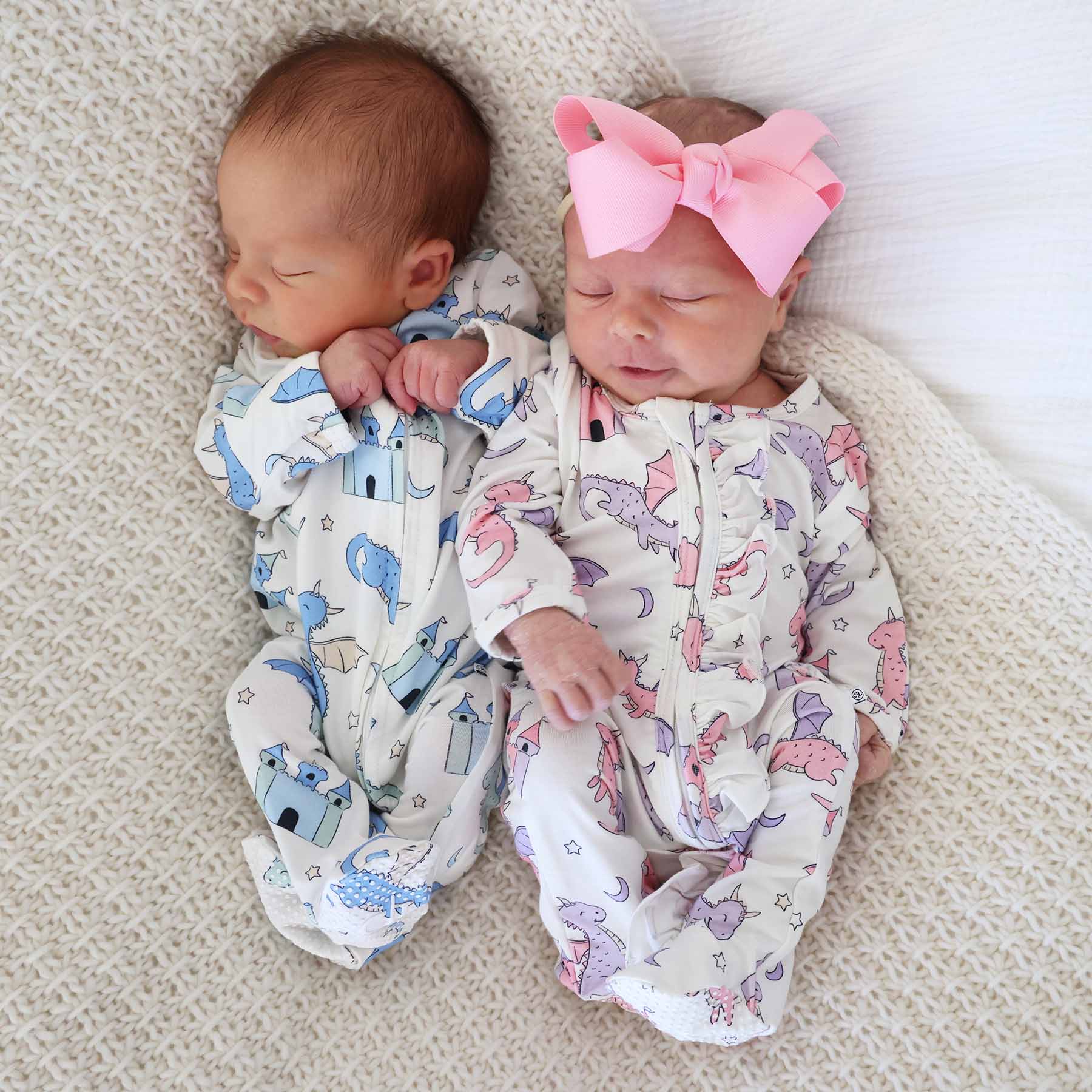 Two newborn babies in patterned pajamas sleeping side-by-side on a knit blanket, one with a pink bow.