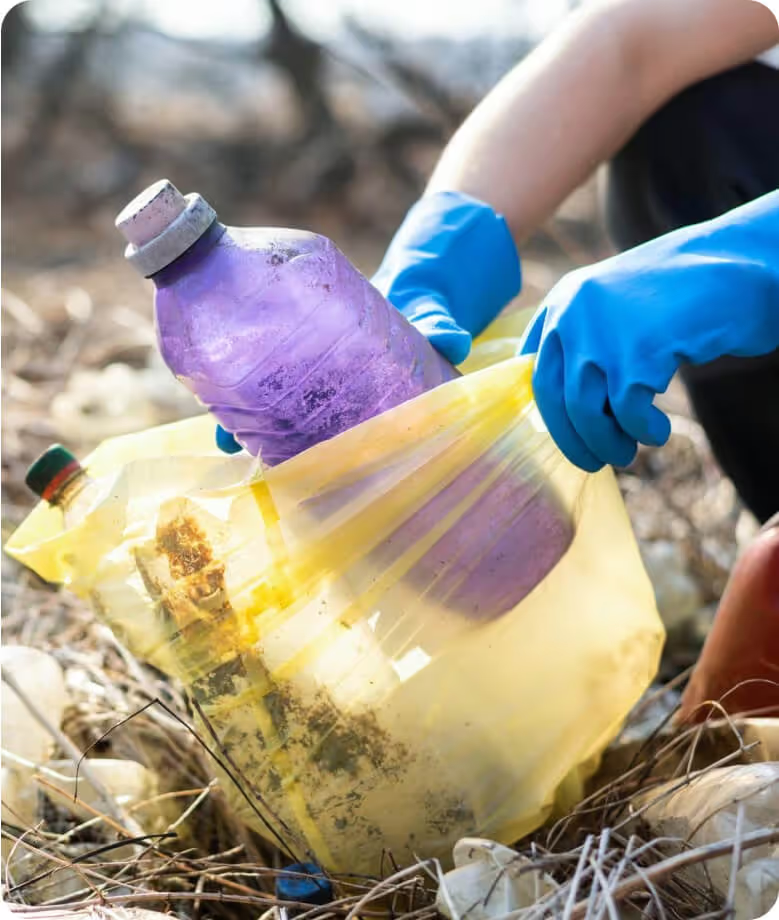 Person with gloves collecting trash in a yellow bag outdoors.