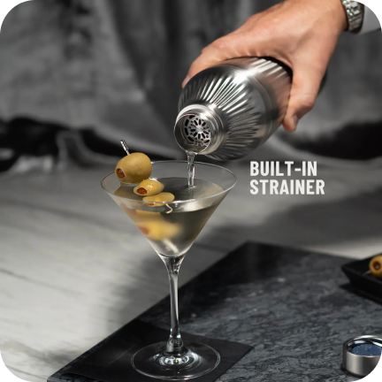 Hand pouring martini from shaker with built-in strainer into glass with olives.