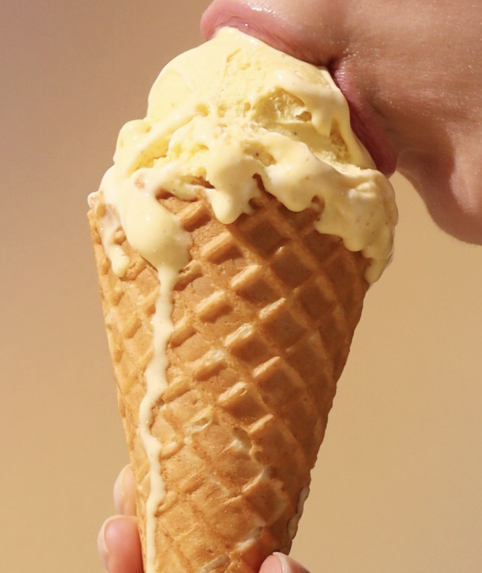 A close-up of a person licking a melting scoop of vanilla ice cream in a waffle cone.