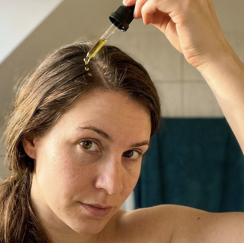 A woman looks at the camera while applying oil to her scalp with a glass dropper.