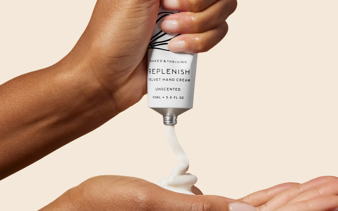 A hand squeezes a tube of Replenish Velvet Hand Cream onto the back of another hand.