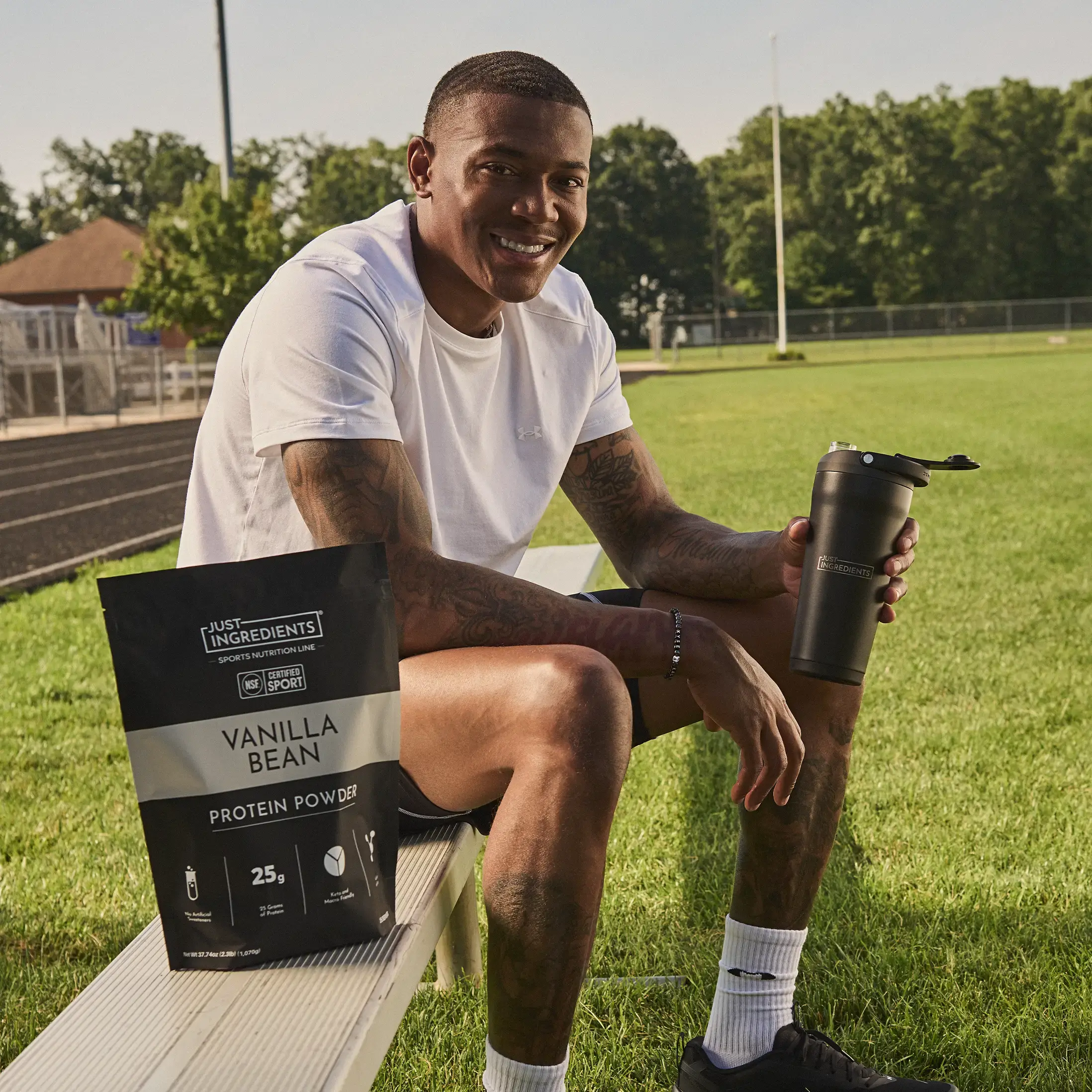 A smiling man sits on a bench at a sports field with protein powder and a shaker bottle.