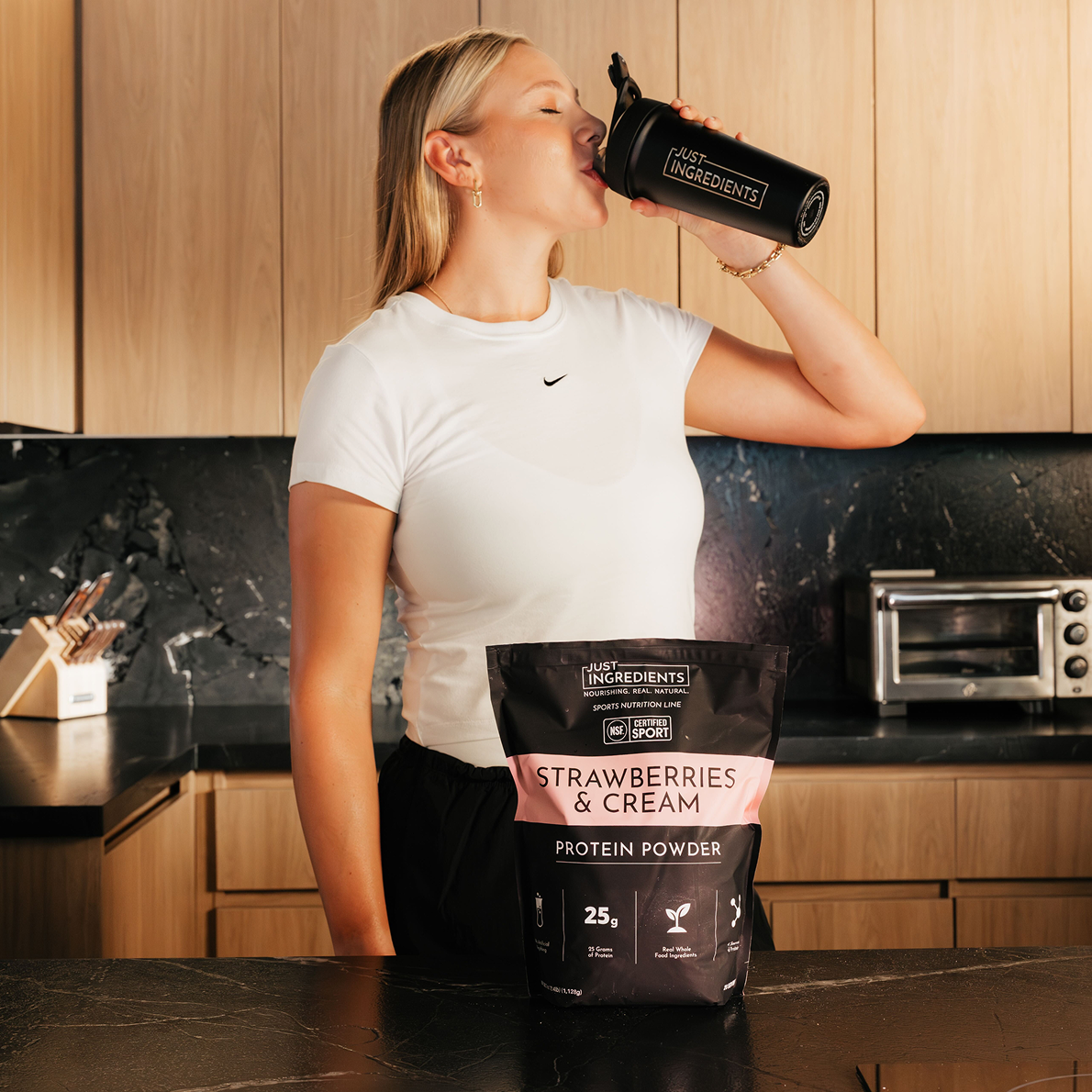 A woman drinks from a black shaker bottle next to a bag of protein powder in a kitchen.