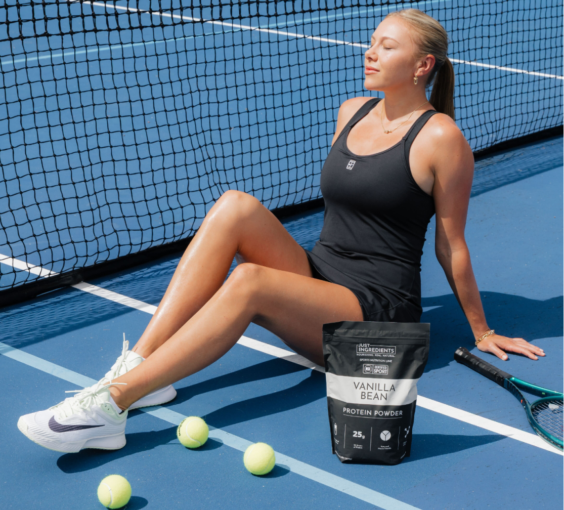 A woman in tennis clothes sits on a blue court next to a bag of protein powder.