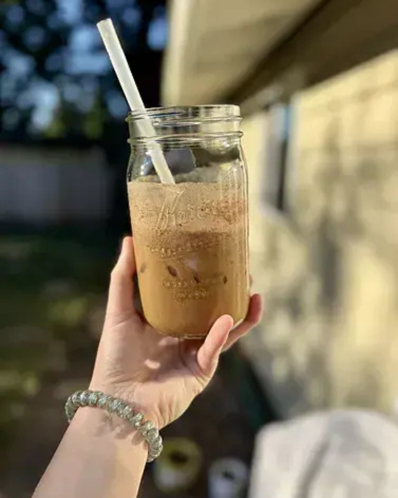 Hand holding a mason jar with iced coffee and straw against outdoor background.