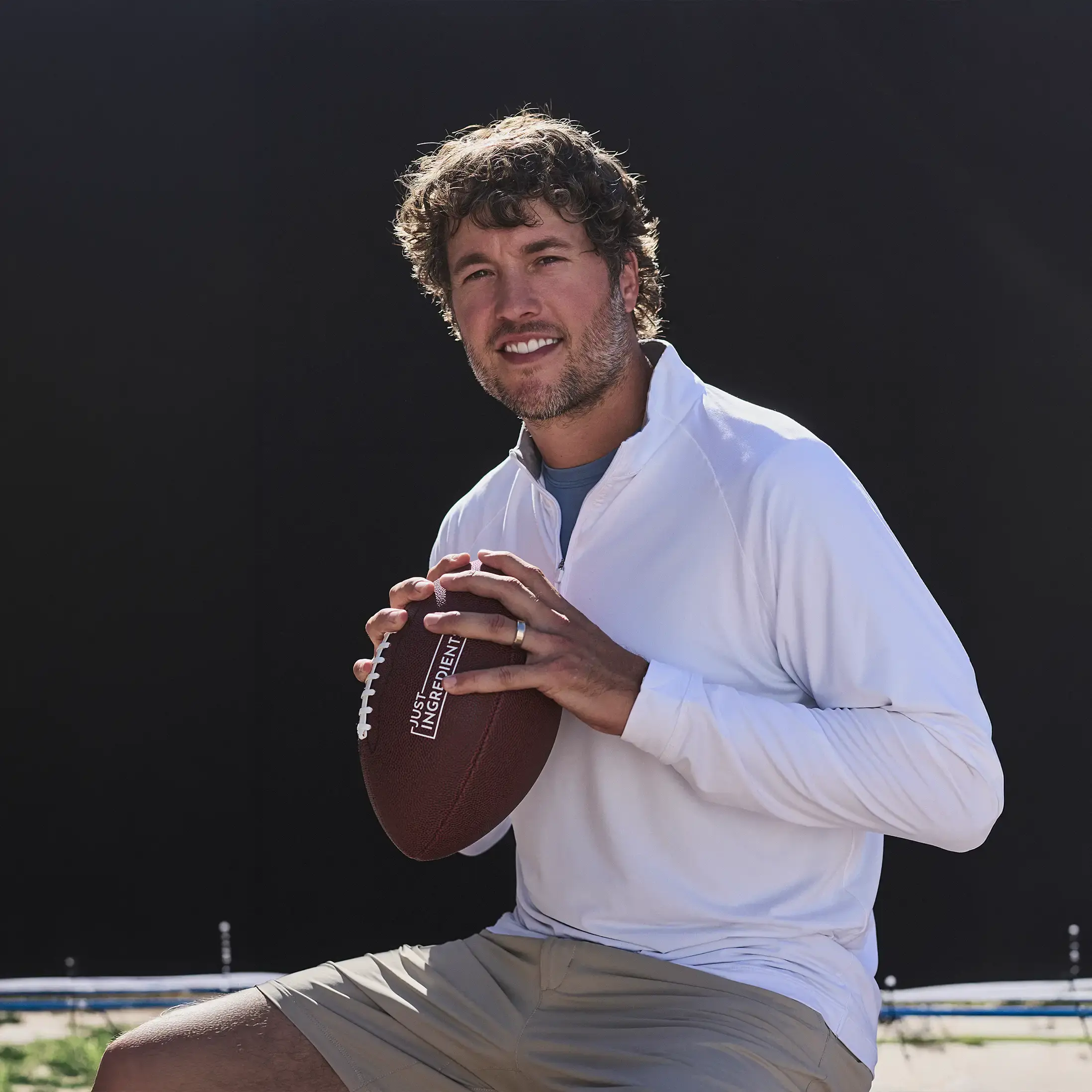 A smiling man with curly hair in a white long-sleeved shirt holds a football against a dark backdrop.