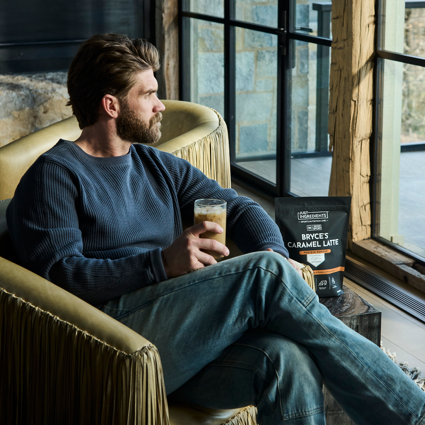 A man in a blue sweater sits in an armchair holding an iced coffee next to a window.
