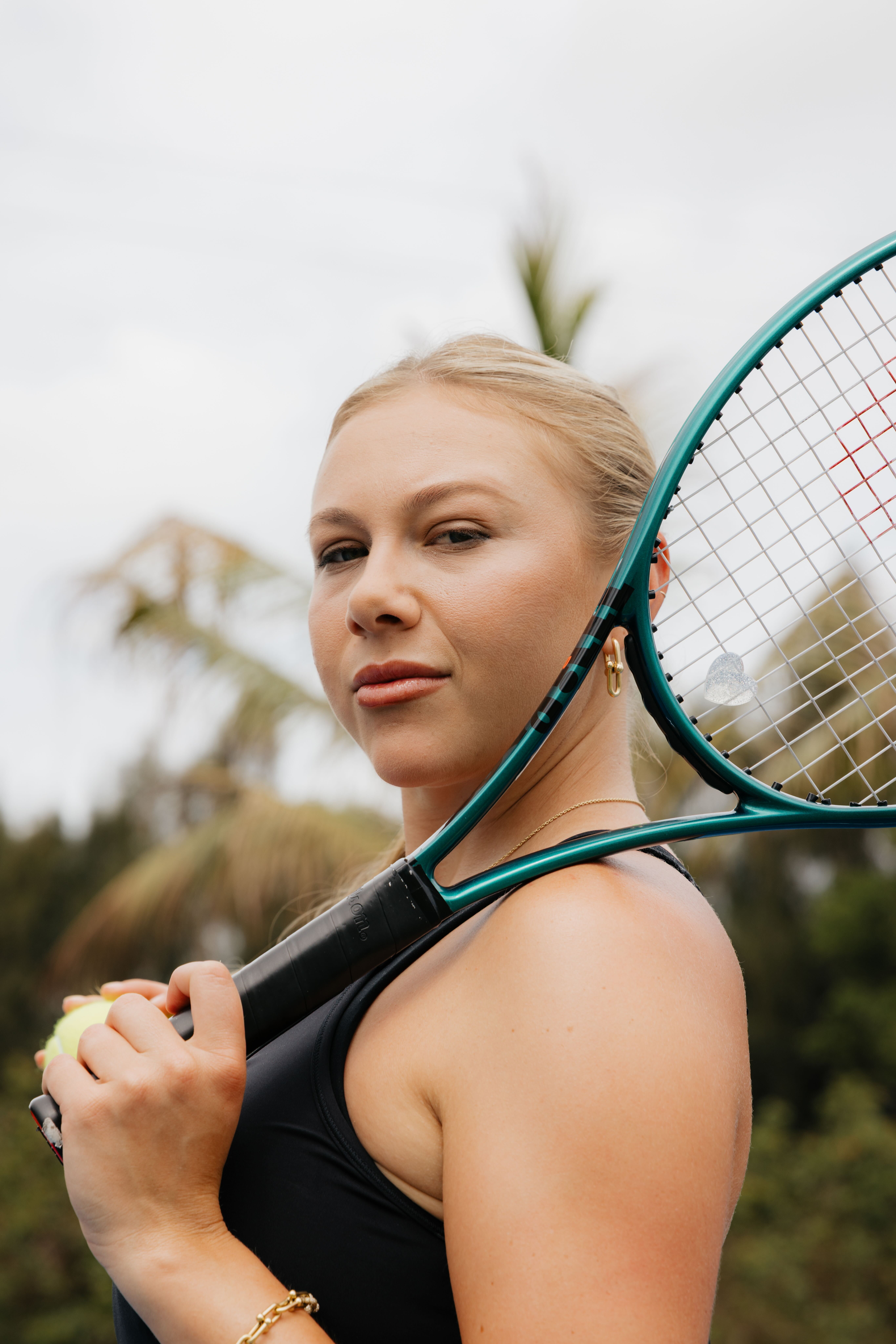 A blonde woman looks at the camera while holding a tennis racket over her shoulder.
