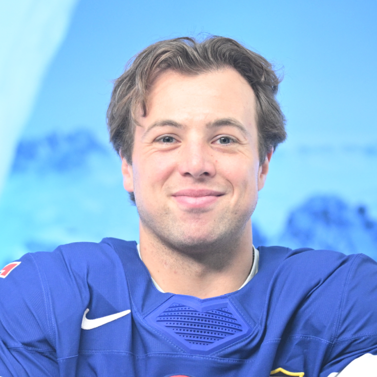 A smiling man with brown hair wearing a blue athletic jersey in front of a blurry blue background.