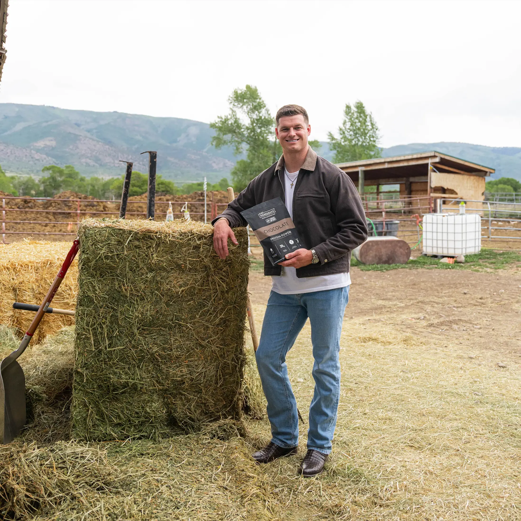 A man on a farm holds a bag of chocolate protein powder while leaning on a hay bale.
