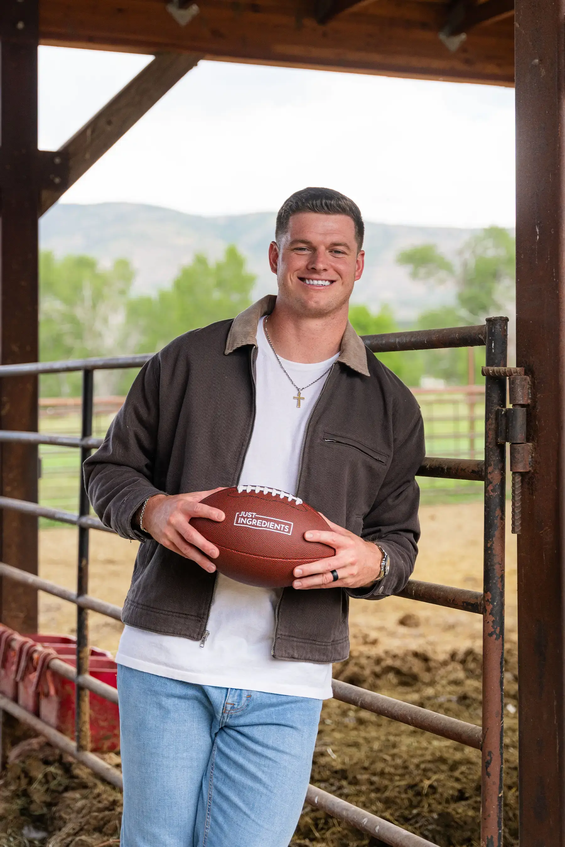A smiling man in a brown jacket and jeans holds a football while standing in a barn.