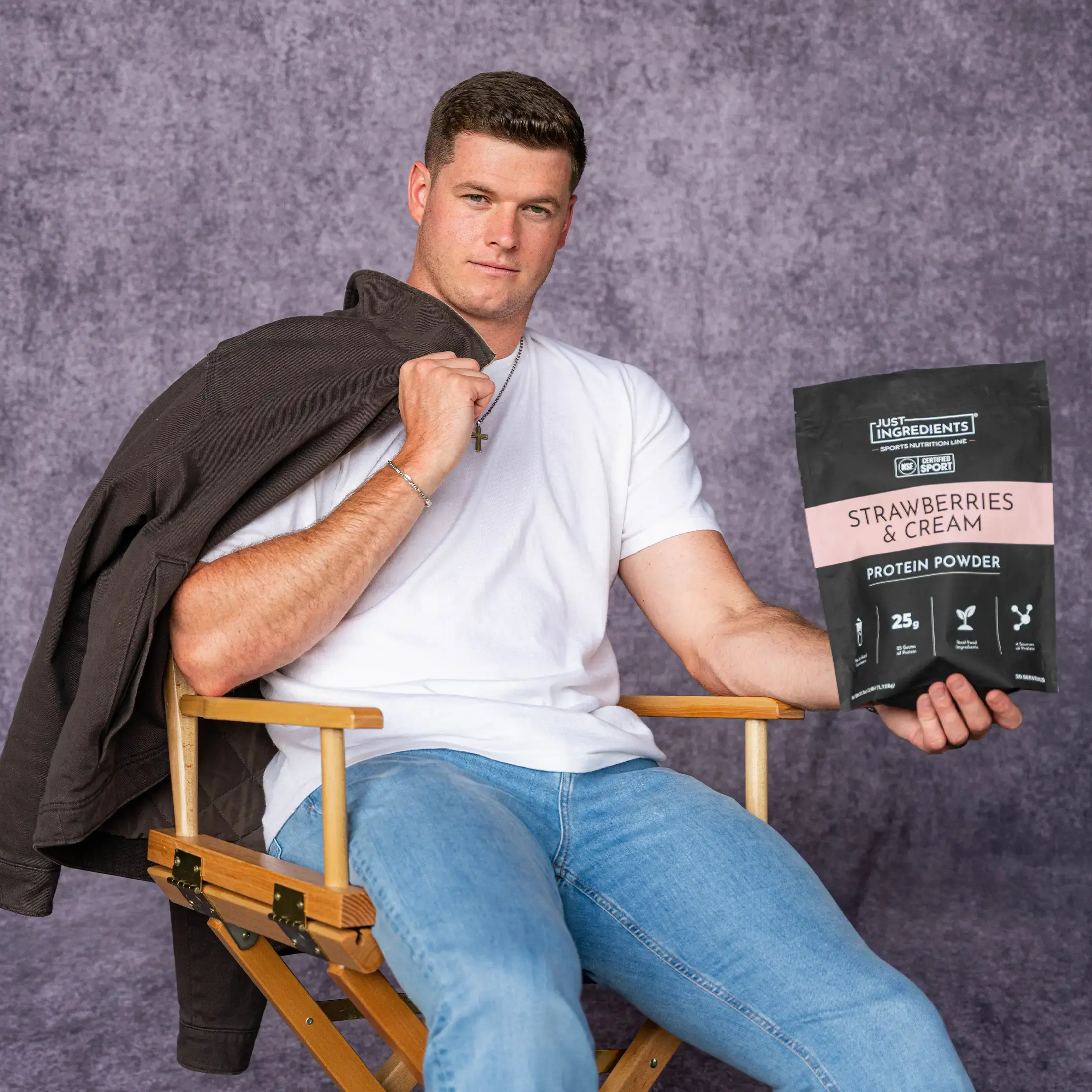 A man in a director's chair holds a bag of Just Ingredients Strawberries & Cream protein powder.