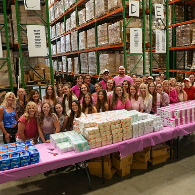 Group of people in a warehouse beside tables of stacked boxed items.