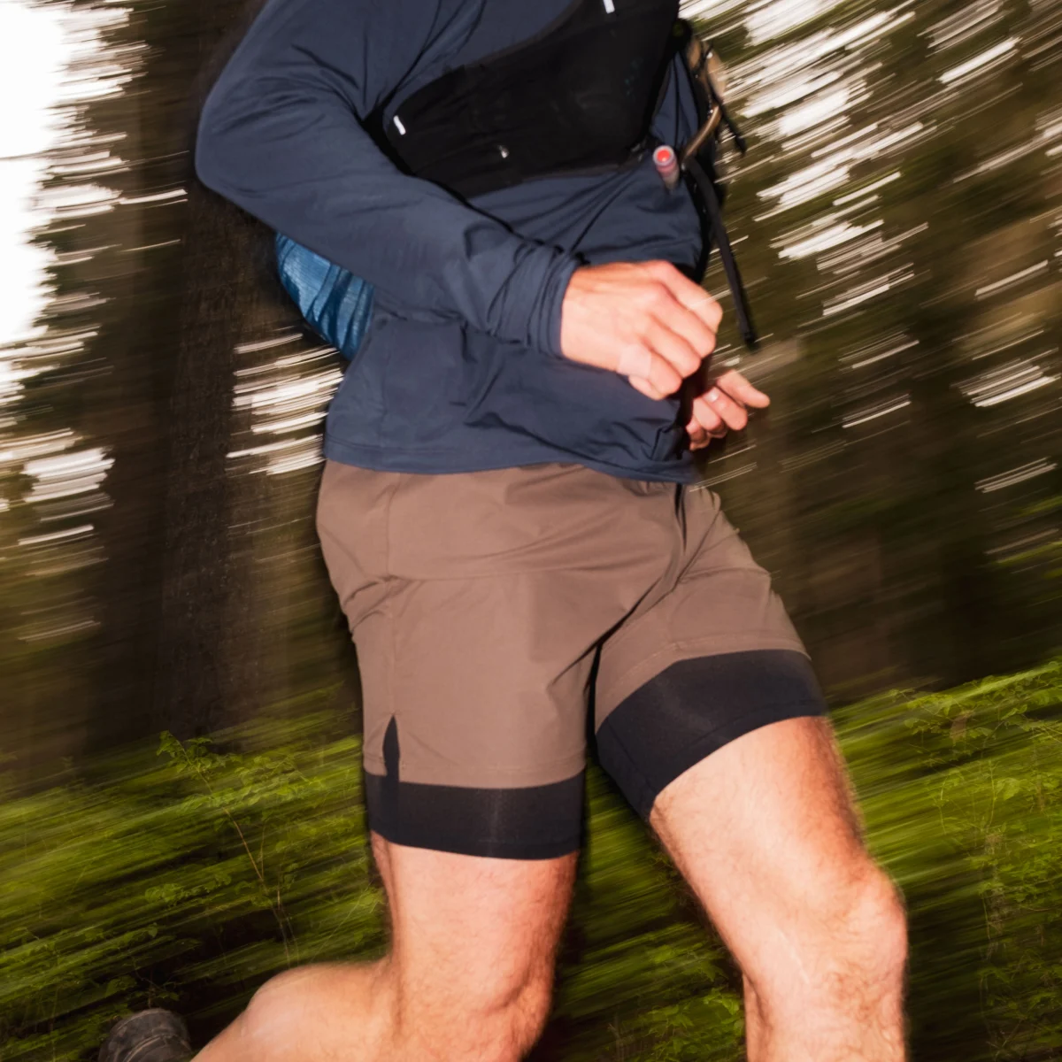 A motion-blurred shot of a person running through a forest wearing athletic shorts and a hydration vest.